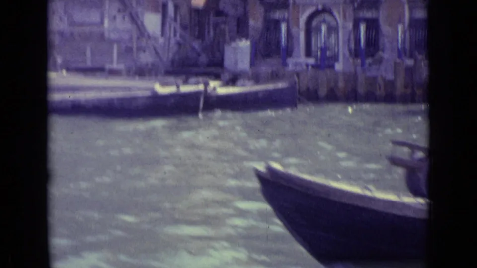 Venice canal view from a moving boat, with a bow in the foreground, moored boat, rippling water, and waterfront architecture.