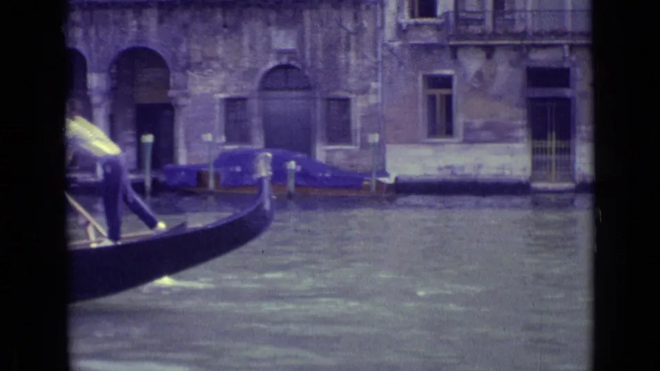 1962 Venice 8mm frame showing a gondola moving across canal water in front of weathered stone buildings and arched openings.