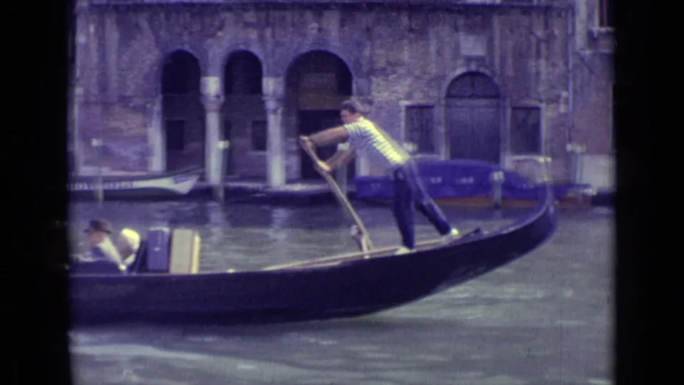 1962 Venice frame of a striped-shirt gondolier rowing passengers past canal-side stone facades and arched doorways.