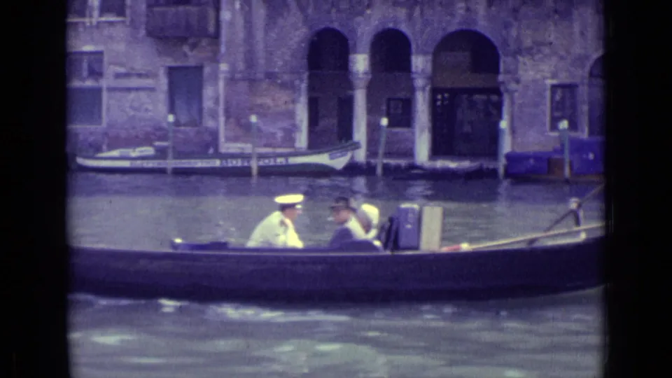 1962 Venice 8mm still showing gondola passengers, including a man in a white hat, moving past weathered canal architecture.