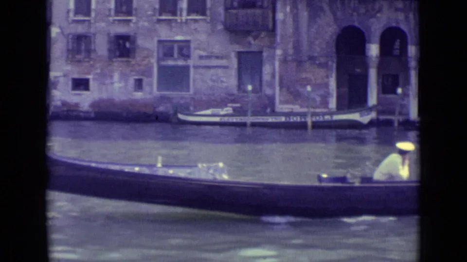 1962 Venice frame with a gondola crossing the foreground, canal water, moored boats, and aged building facades behind it.