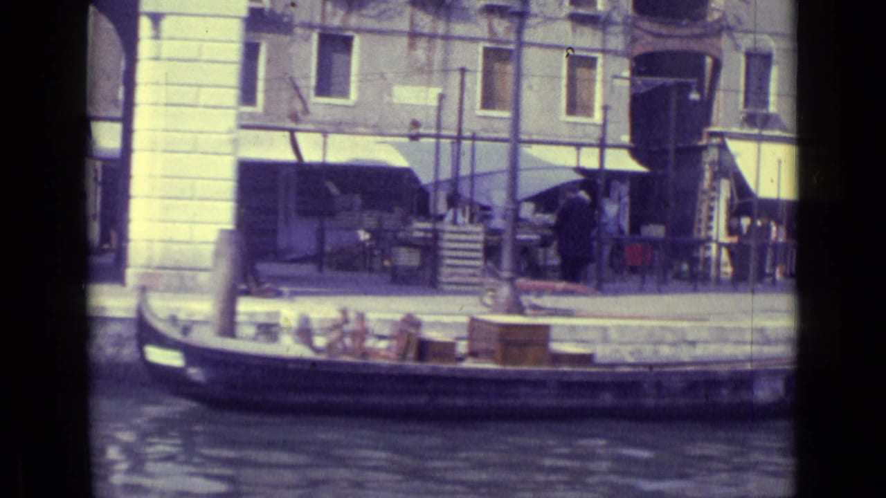 Gondola passes a waterfront building with awnings and pedestrians near the canal.