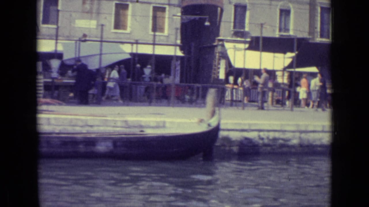 Gondola bow and canal-side pedestrians appear in front of a building with awnings.