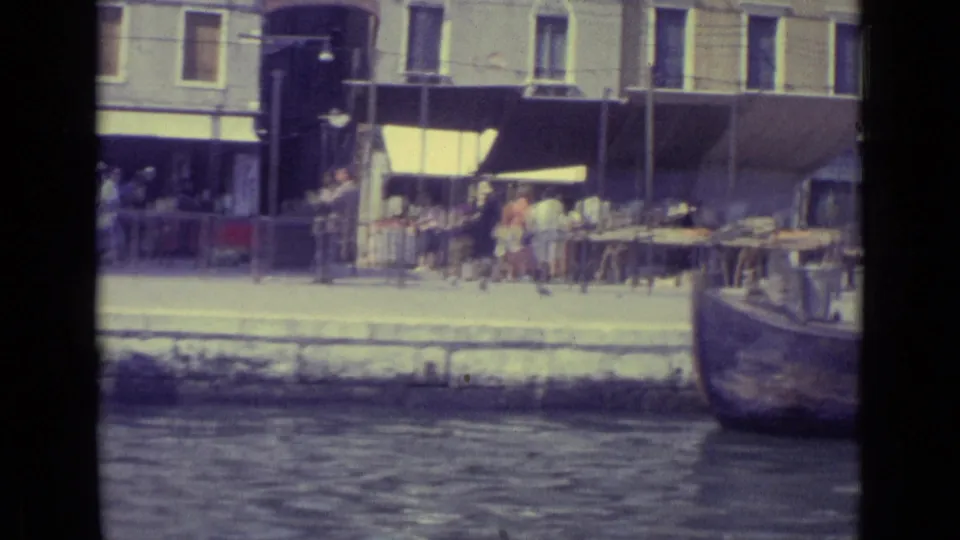 1962 Venice home-movie frame with market-style tables, awnings, pedestrians, stone facades, and canal water in the foreground.