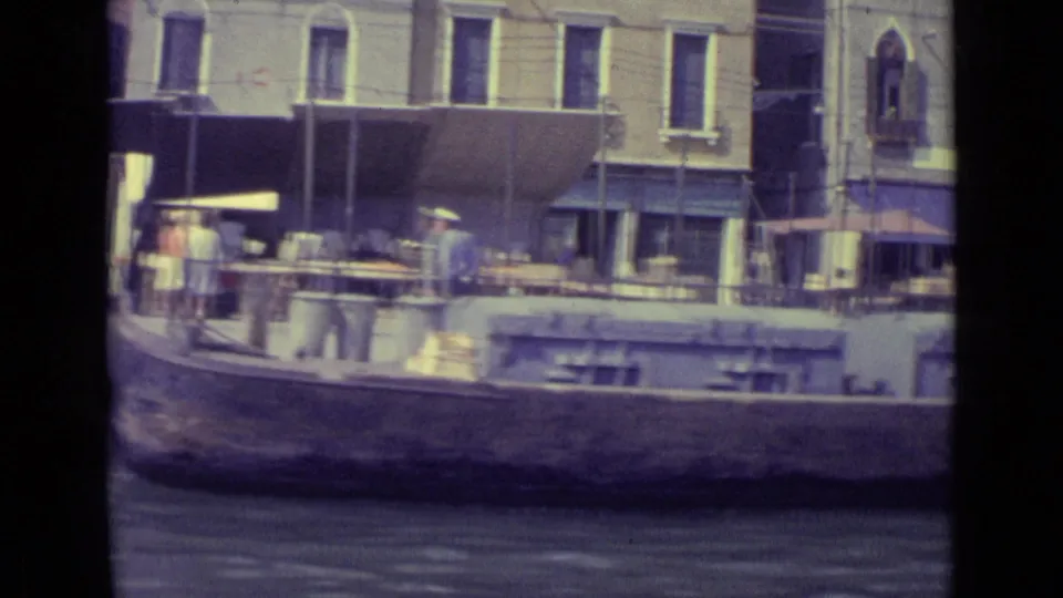 1962 Venice canal footage showing a passing boat edge, pedestrians, waterfront awnings, building windows, and a quayside umbrella.