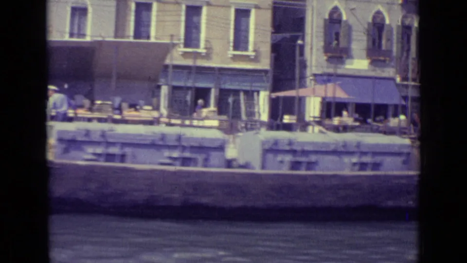 1962 Venice canal view with shuttered buildings, blue awnings, outdoor cafe seating, and a moored boat along the embankment.