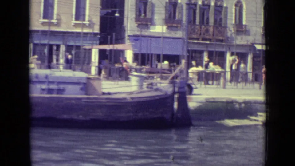 1962 Venice waterfront with cafe awnings, pedestrians, arched windows, and a boat moored along the canal edge.