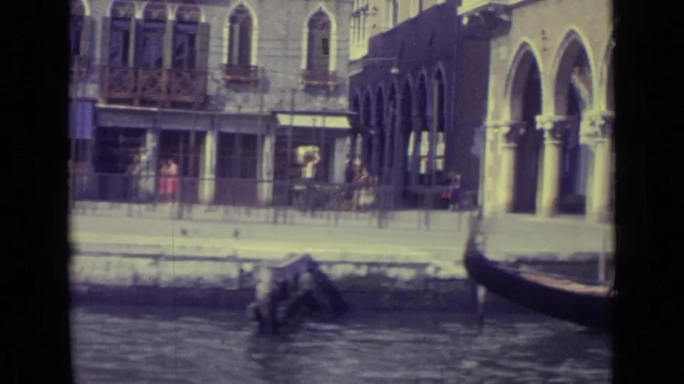 1962 Venice canal view with a gondola bow, arched arcade, cafe awnings, pedestrians, and rippled water in the foreground.