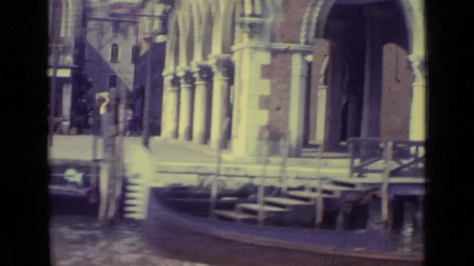 1962 Venice frame with canal water, a long boat, stone steps, arched waterfront architecture, and sunlit building facades.