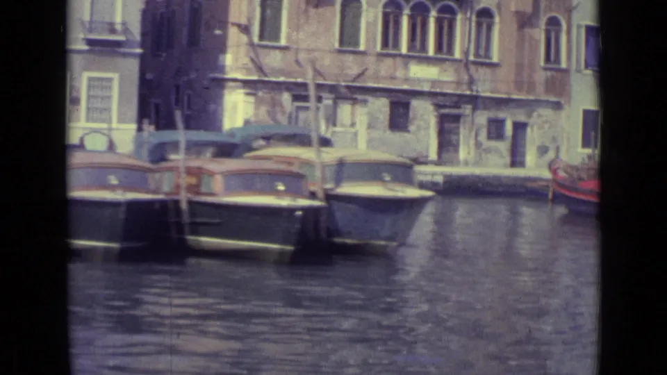 1962 Venice frame showing moored boats, canal water, vertical mooring poles, arched windows, and sunlit stone buildings.