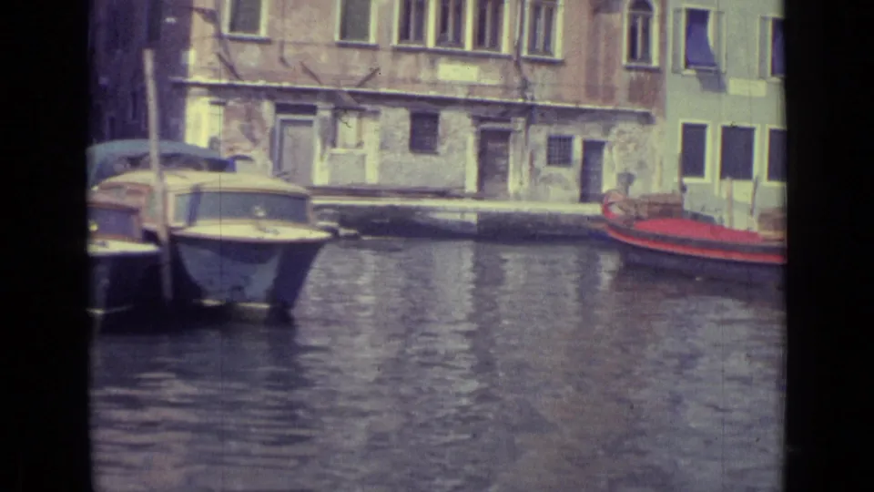 1962 Venice home-movie frame showing small boats tied along a canal with worn building facades behind them.