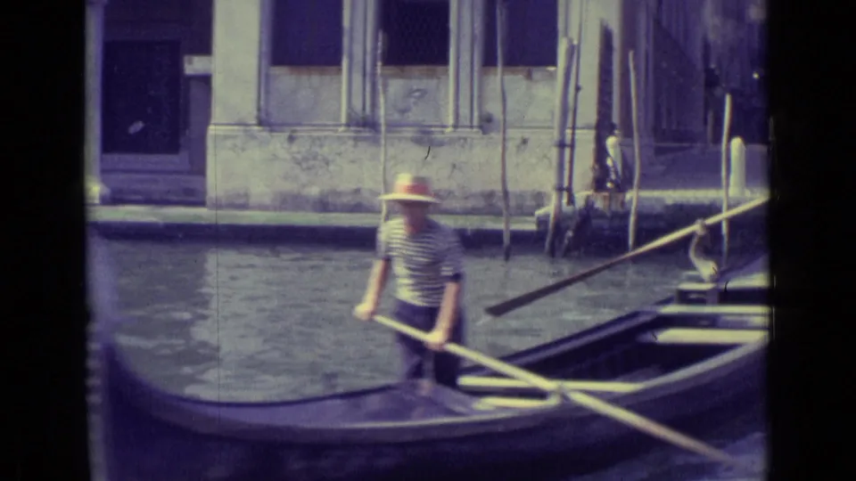 1962 Venice frame with a gondolier in a striped shirt and straw hat steering a gondola near canal-side buildings.