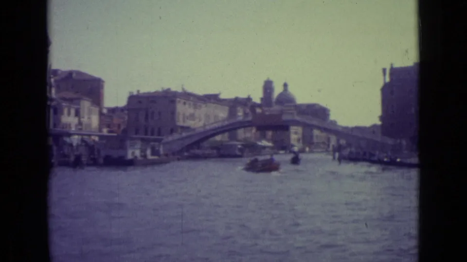 Venice canal traffic passes beneath the Rialto Bridge in a faded 1962 home-movie frame with historic facades along the water.