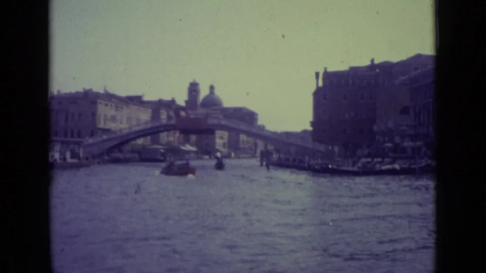 1962 Venice home-movie frame looking along the Grand Canal toward the Rialto Bridge, with boats on the water and dense waterfront architecture.