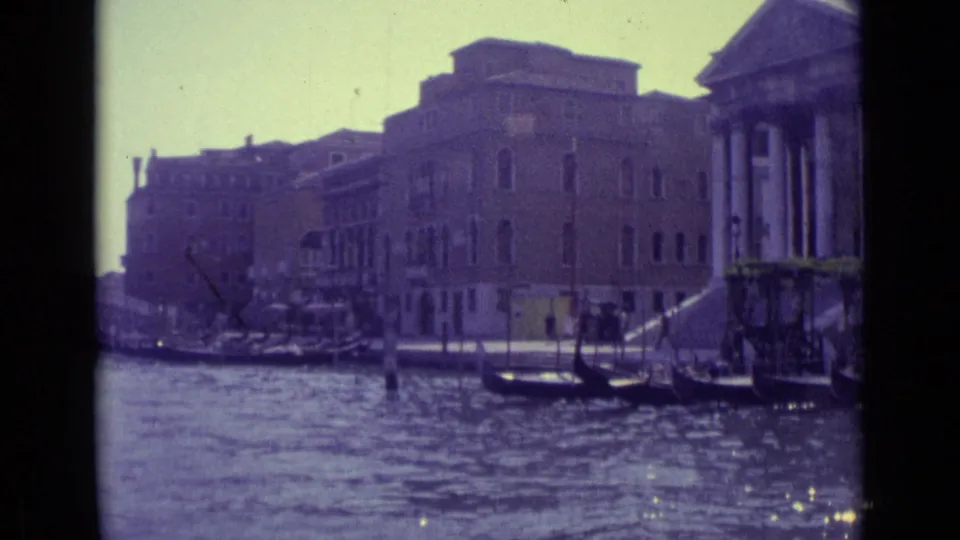 1962 Venice canal view showing moored gondolas, Venetian buildings, and a prominent classical-style facade with columns at the right edge.