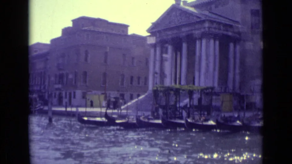 1962 8mm Venice frame with gondolas lined along the canal, stone waterfront steps, and a large classical-style building with columns.