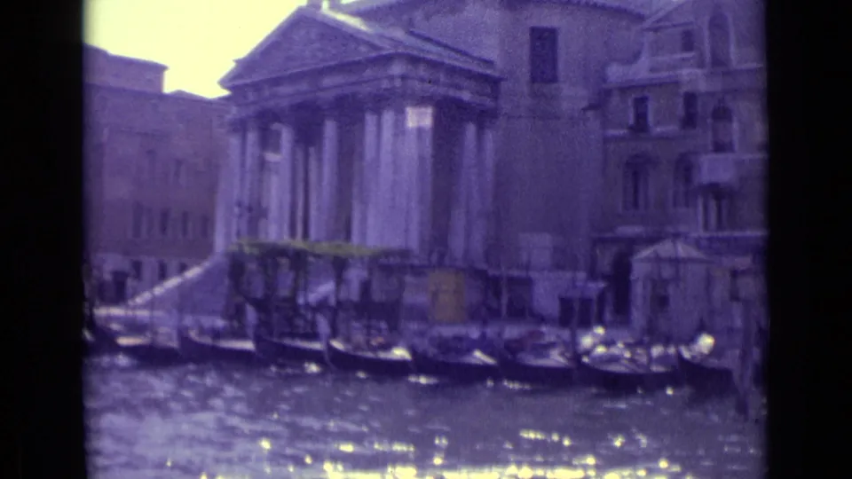 1962 Venice home-movie frame with moored gondolas, bright canal reflections, waterfront steps, and classical columns beside older buildings.