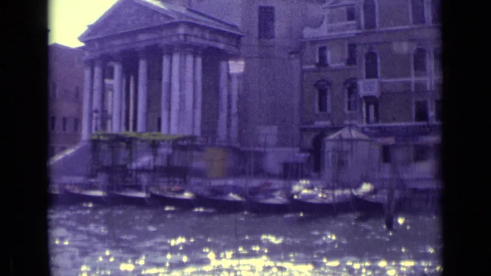 1962 Venice home-movie frame showing a canal, moored boats, bright water reflections, and a classical columned structure near historic buildings.