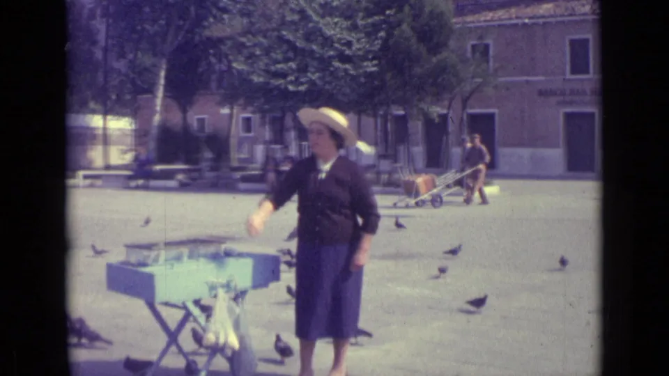 1962 Venice frame showing a woman in a dark dress and straw hat feeding pigeons near a blue table in a public square.
