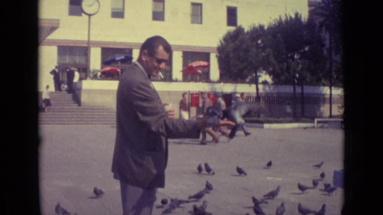 Man in a suit smokes while holding out food for pigeons in a public square.