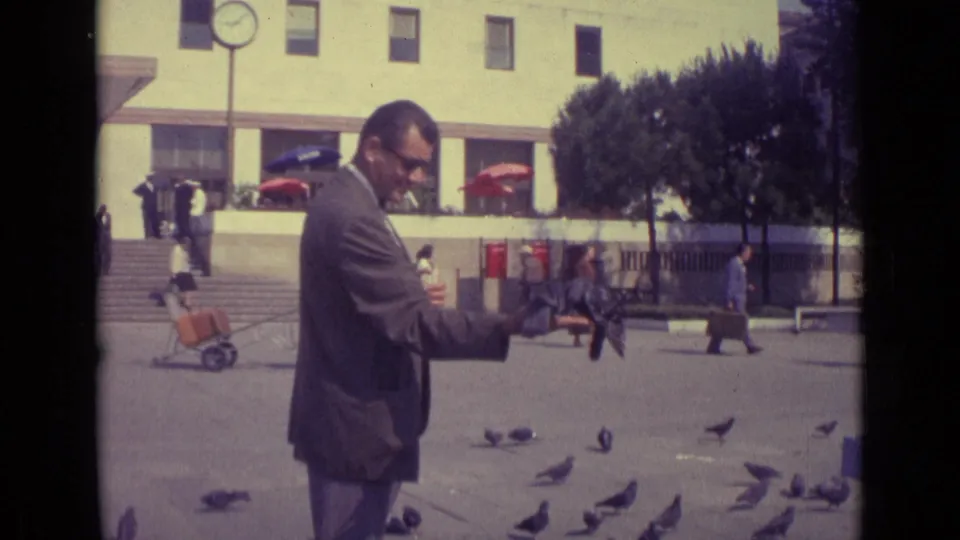 The man continues feeding pigeons in the Venice square, with a cigarette in his mouth and people walking behind him.