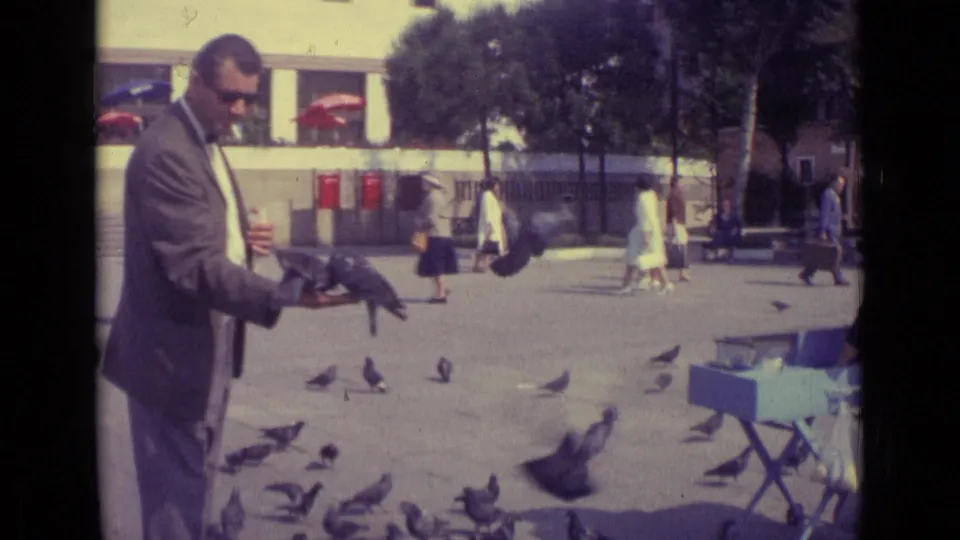 In the final frame, pigeons move around the suited man in the Venice square while pedestrians and red umbrellas remain visible behind him.