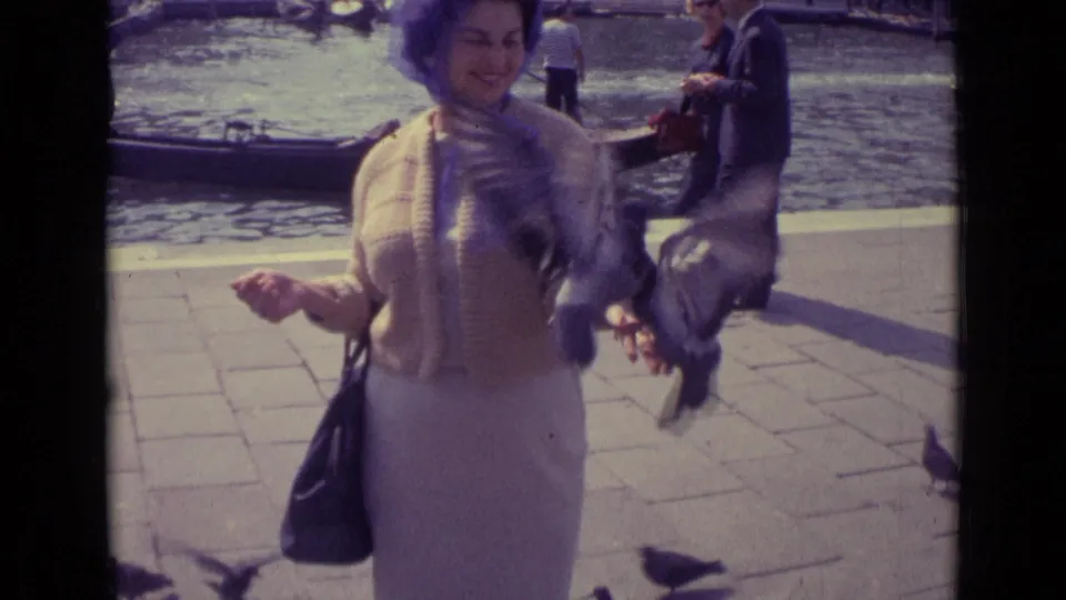 In a 1962 Venice canal scene, a smiling woman feeds pigeons at close range while gondolas and visitors remain visible behind her.