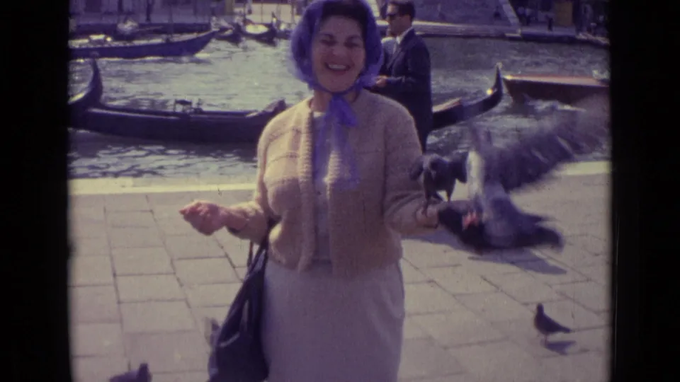 The 1962 Venice home-movie frame shows a woman laughing as a pigeon flies near her hand, with canal water and gondolas behind her.
