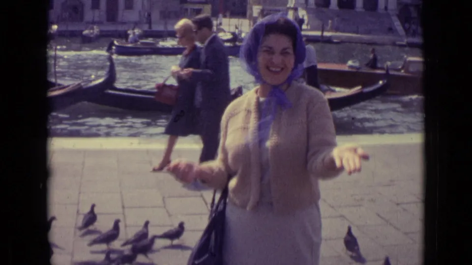 At the end of the 1962 Venice clip, the woman stands with open hands while pigeons gather on the canal walkway and gondolas sit behind her.