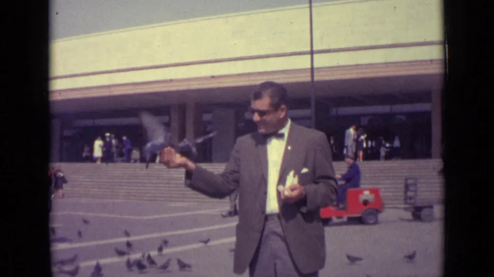 In Venice, Italy, the suited man continues feeding pigeons in the plaza, with pedestrians, steps, and a small red cart in the background.