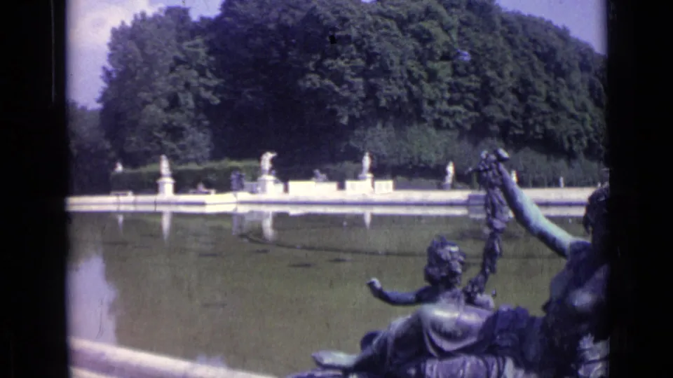 1962 Versailles home-movie frame showing a formal pool, distant statues, dense trees, and a foreground bronze sculpture by the water.