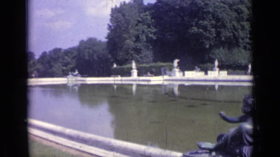 1962 Versailles garden view across a long reflecting pool toward white statues, stone edging, and dense trees.