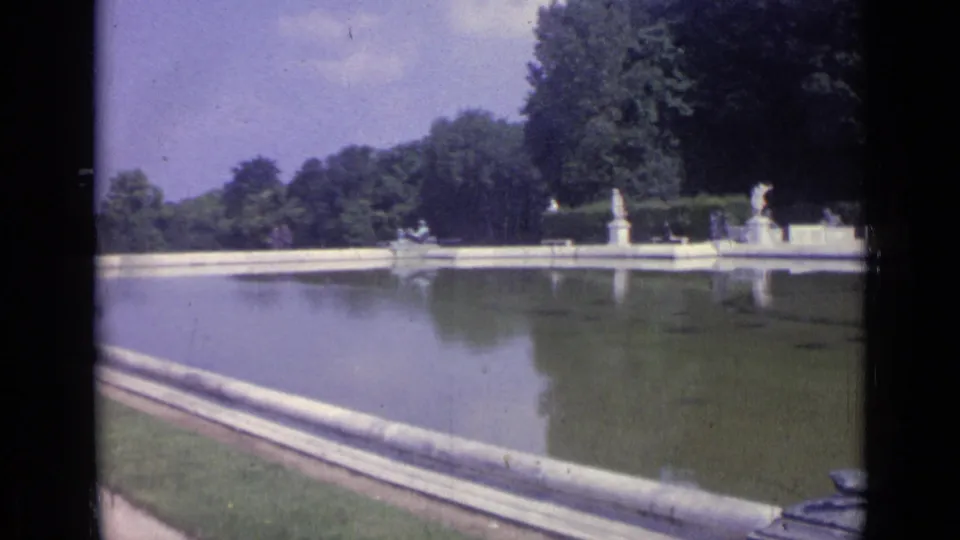 1962 Versailles footage frame showing the pool surface, formal stone edging, distant garden statues, and surrounding trees.