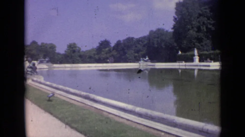 1962 Versailles home-movie frame with a wide pool view, formal statues, tree-lined background, walkway, and reflected stonework.