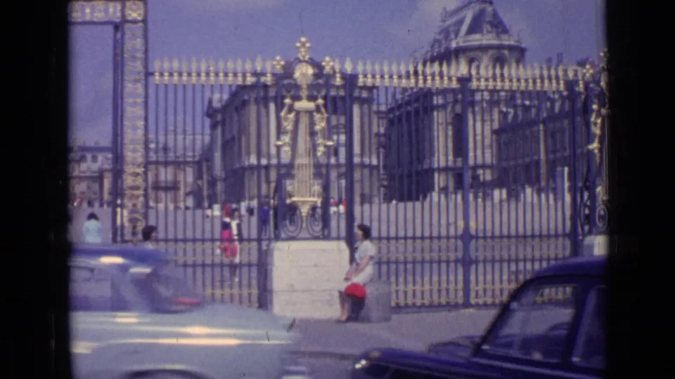 1962 8mm view of the Palace of Versailles entrance, with ornate iron gates, tourists, and a light-colored car crossing the foreground.
