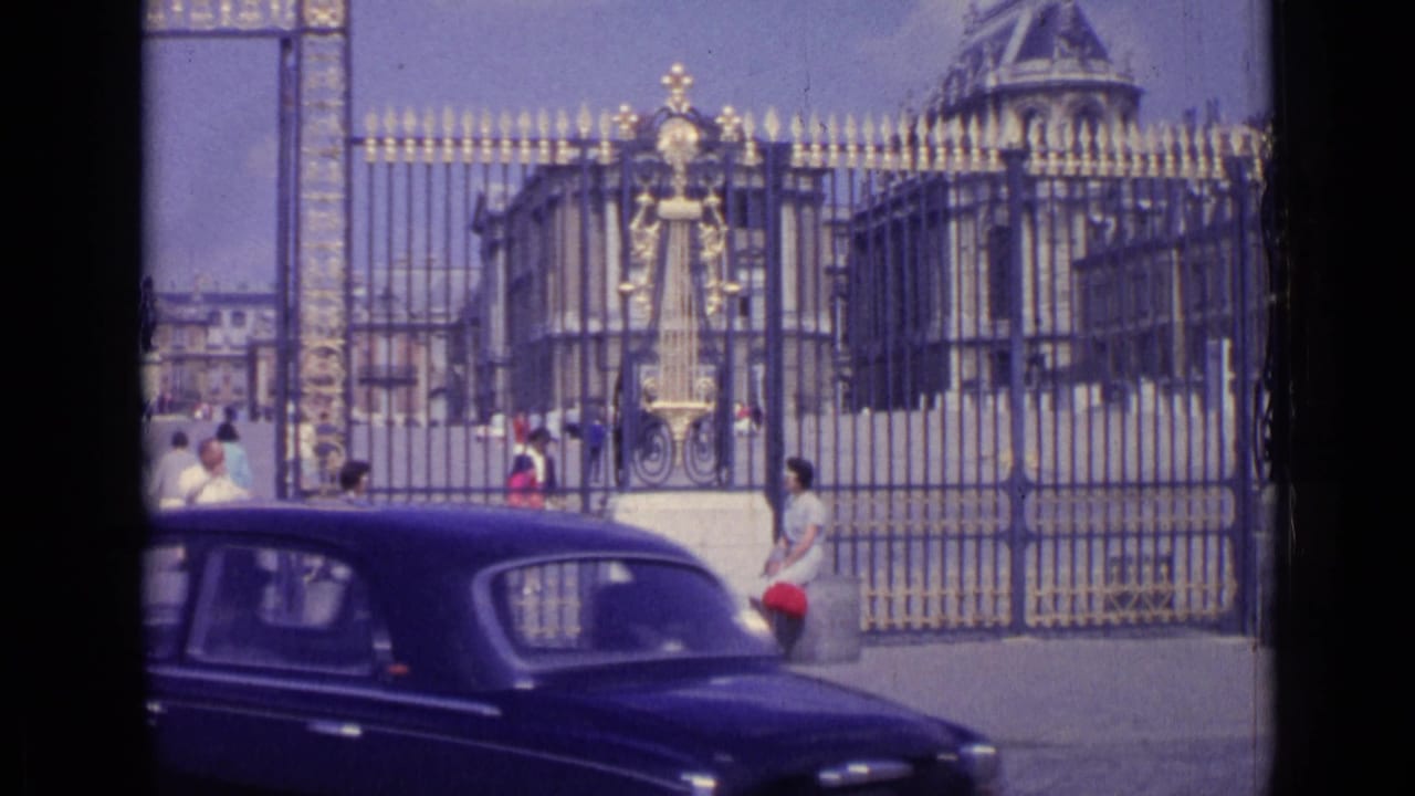 Dark classic car passing in front of the Versailles gates with visitors behind it.