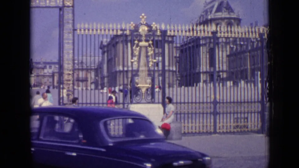 A dark early-1960s car moves across the foreground at the Versailles entrance while visitors stand and sit near the gates.
