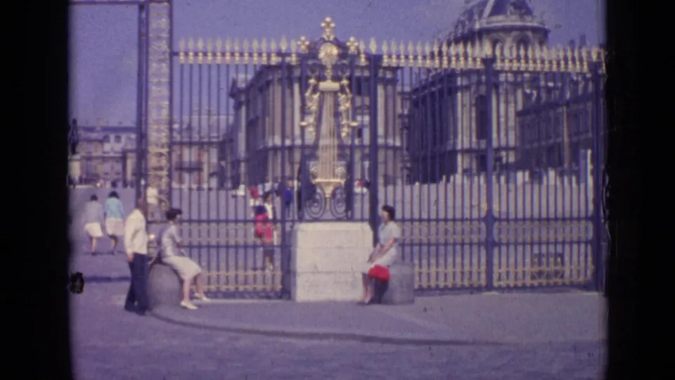 Visitors in period clothing gather near the Palace of Versailles entrance, with the ornate gates and palace buildings behind them.