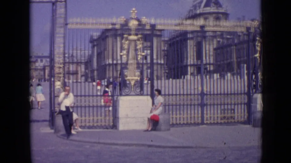A man walks through the foreground while visitors sit near the ornate Palace of Versailles entrance gates in 1962 home-movie footage.
