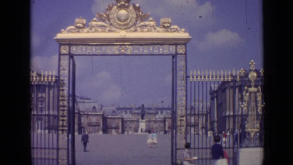 1962 8mm view of the Palace of Versailles entrance gate, with gilded ironwork, open courtyard, and tourists walking beyond.