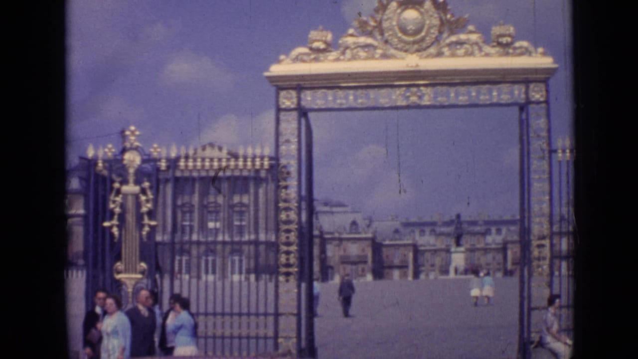 1962 home-movie frame at Versailles showing visitors near the gilded entrance gate and the palace facade across the courtyard.