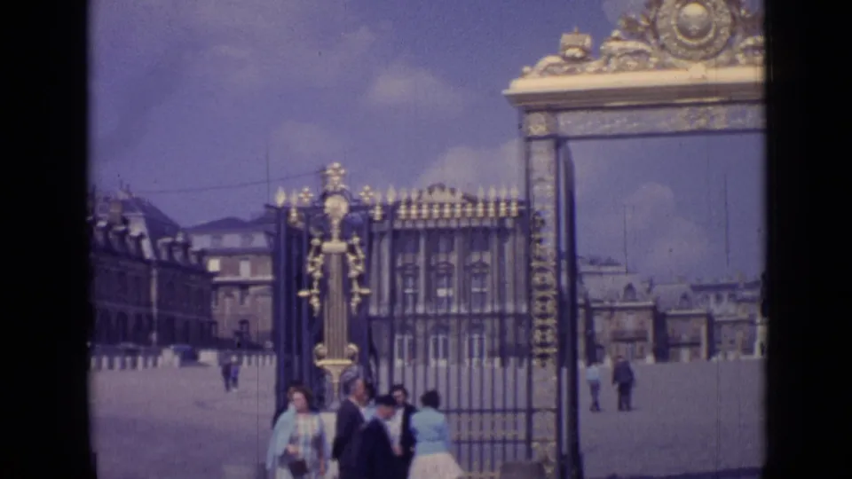 1962 Versailles 8mm frame with the camera angled across the ornate gate, iron fence, courtyard, and several tourists in period dress.