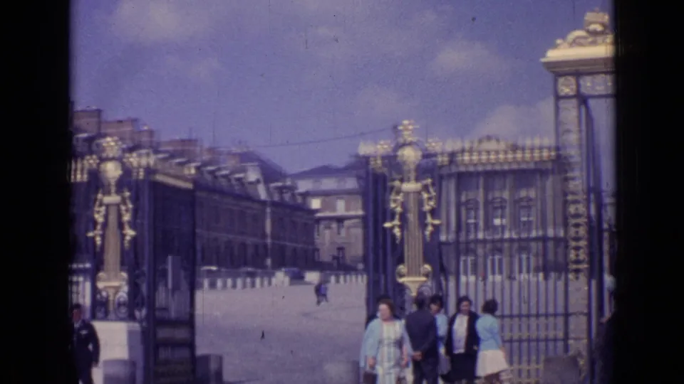 1962 8mm frame of tourists passing the Versailles entrance gate, with iron fencing, palace buildings, and open courtyard behind them.