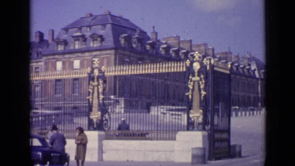 The Versailles gate is seen from the street side, with visitors near the stone base and part of a dark car entering the frame.