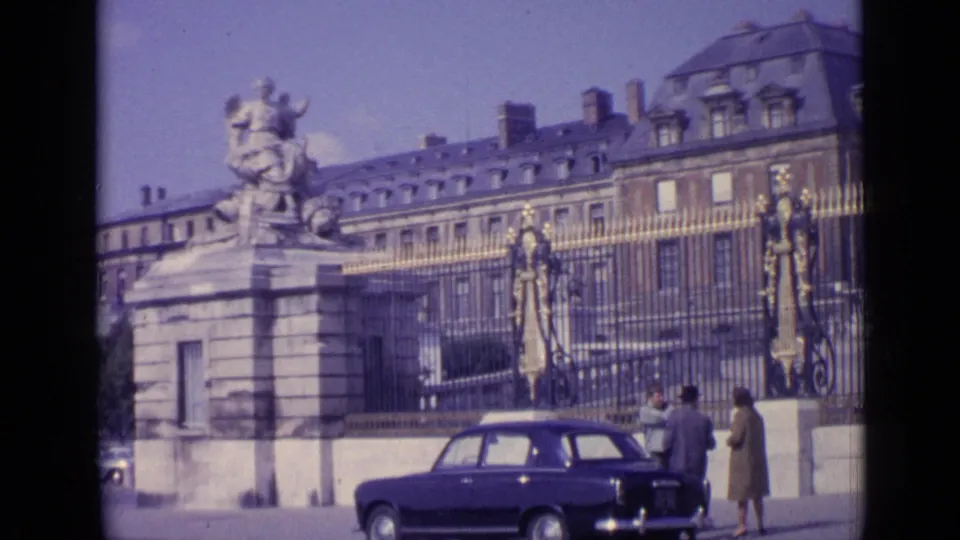 A street-level 1962 view of Versailles shows a large stone sculpture, gilded entrance gates, pedestrians, and a dark car in front.