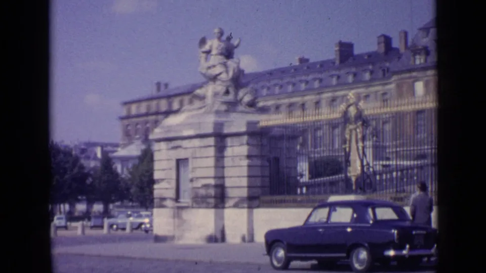 Versailles street view from 1962 with a prominent stone sculpture, palace fencing, distant traffic, and a dark car near the curb.