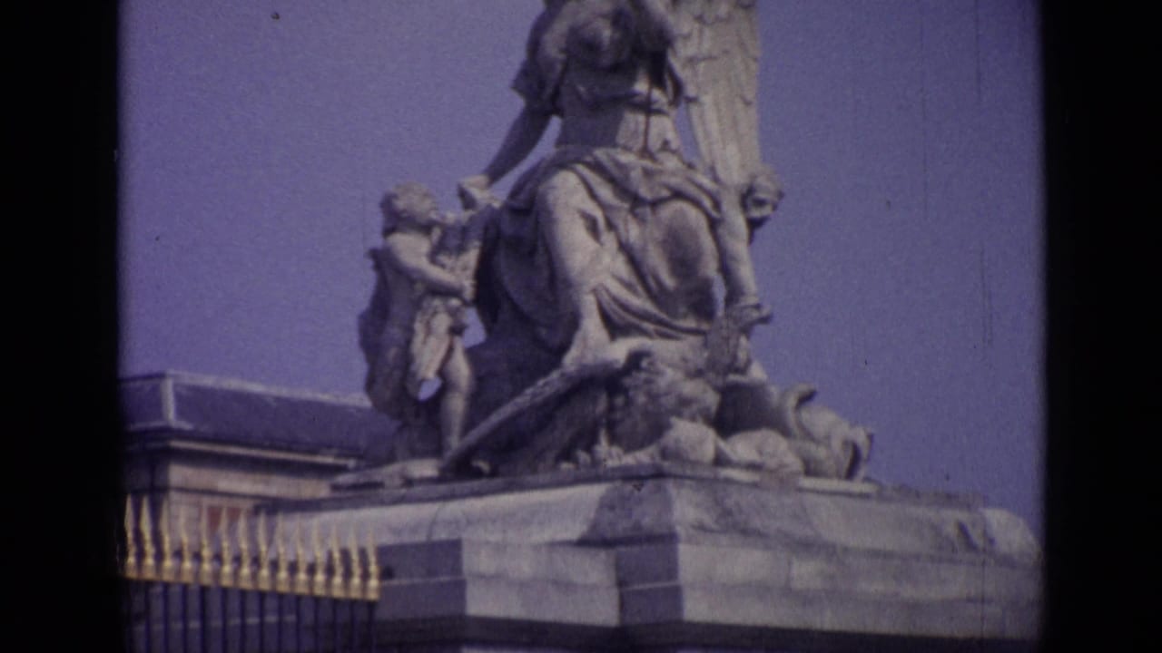 Close view of a stone monument with winged figures and a palace roof edge behind it.