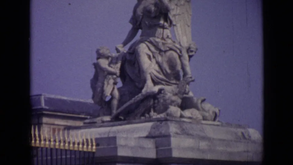 1962 8mm view at Versailles showing part of a stone sculptural group with winged figures, a railing, and palace architecture.