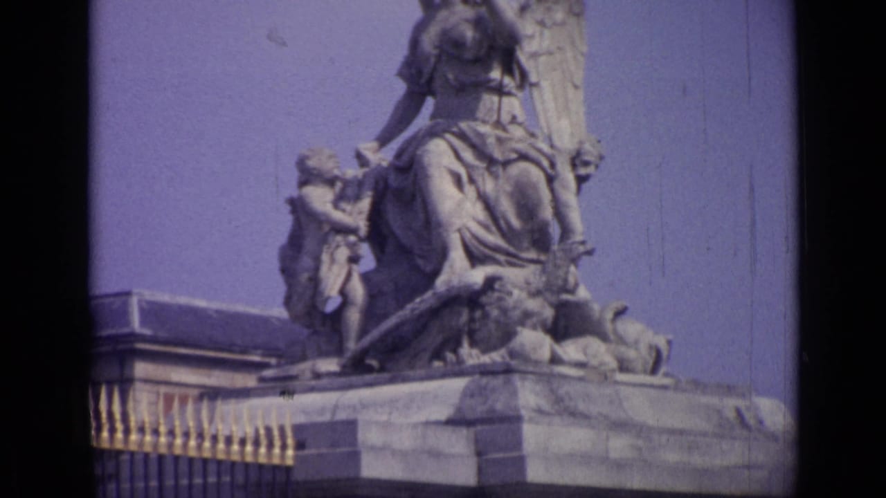 Stone monument detail with a winged figure, smaller figures, railing, and blue sky.