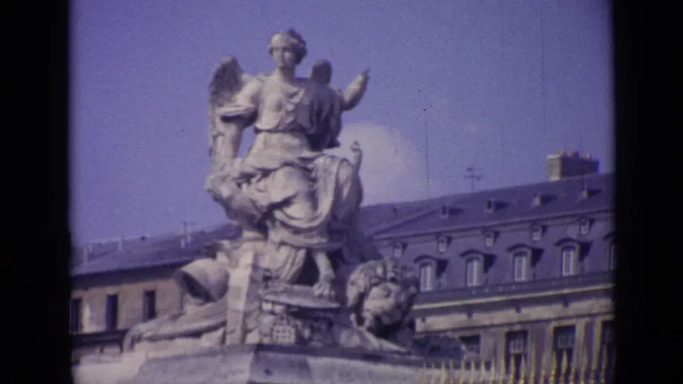 Versailles 8mm frame showing a seated stone figure on a monument, palace roofline, chimneys, railing, and faded sky.